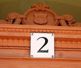 Hausnummer „2“ auf verziertem, terrakottafarbenem Holzzierrat.