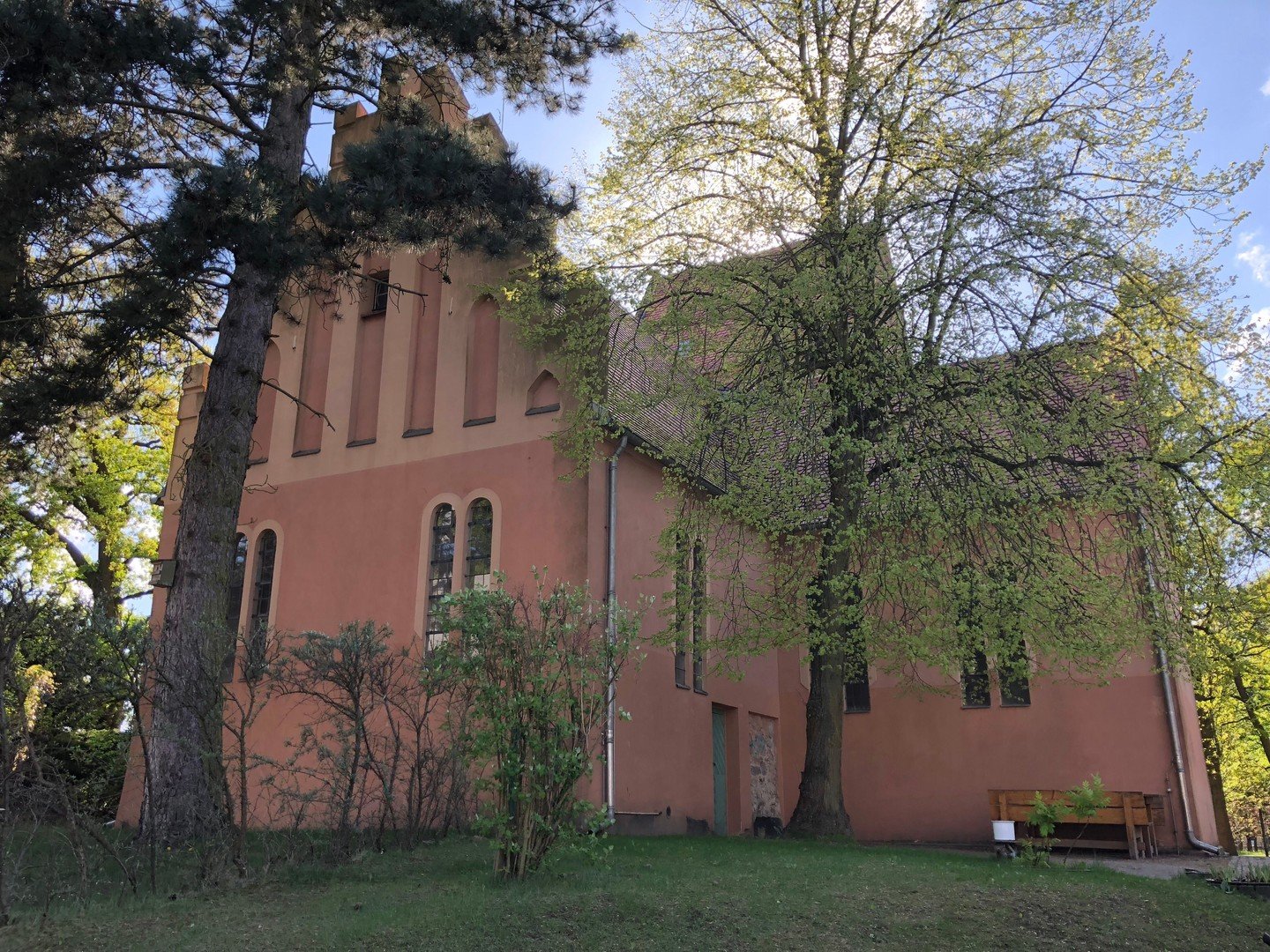Historische, terrakottafarbene Kirche, umrahmt von großen Bäumen.